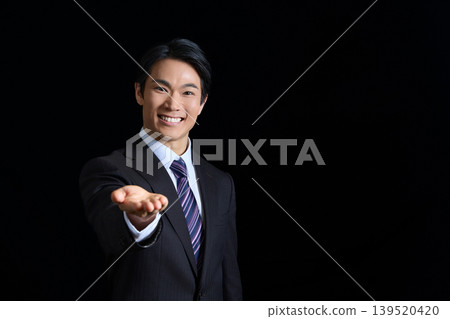 A smiling businessman extending his hand against a black background. A smiling businessman extending his hand against a black background. 139520420