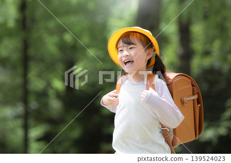 A smiling girl runs to school with her backpack amidst the fresh greenery; a first-grader wearing a yellow hat, her new life, commuting, school, and educational insurance. A smiling girl runs to school with her backpack amidst the fresh greenery; a first-grader wearing a yellow hat, her new life, commuting, school, and educational insurance. 139524023