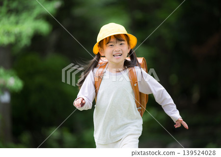 A smiling girl walks to school with her backpack amidst the fresh greenery; a first-grader wearing a yellow hat, her new life, commuting, school, and educational insurance. A smiling girl walks to school with her backpack amidst the fresh greenery; a first-grader wearing a yellow hat, her new life, commuting, school, and educational insurance. 139524028
