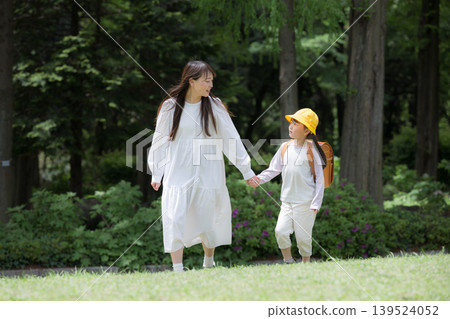A mother and her daughter, wearing school bags, walk hand in hand and gaze into each other's eyes through a park lush with new greenery. A full-body scene of a mother and daughter going to school. A mother and her daughter, wearing school bags, walk hand in hand and gaze into each other's eyes through a park lush with new greenery. A full-body scene of a mother and daughter going to school. 139524052