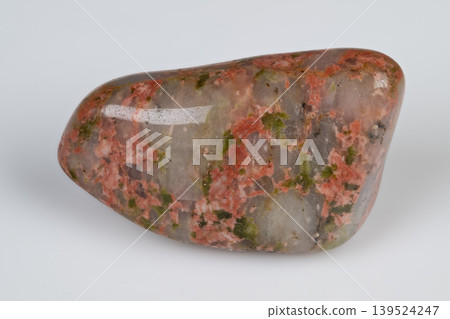 Smooth unakite pebble displaying lively mineral colors on plain surface. Polished unakite gemstone characterized by vibrant mineral inclusions against neutral backdrop 139524247