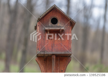 Rustic nest shelter background. Weathered bird shelter amidst moss and winter trees. Aged red birdhouse on wooden post with mossy surroundings and blurred forest backdrop Rustic nest shelter background. Weathered bird shelter amidst moss and winter trees. Aged red birdhouse on wooden post with mossy surroundings and blurred forest backdrop 139525106