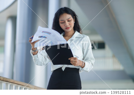 Professional businesswoman checks a smartphone while holding reports in a modern office, showing mobile communication, digital workflow, corporate planning, career success, and productivity. Professional businesswoman checks a smartphone while holding reports in a modern office, showing mobile communication, digital workflow, corporate planning, career success, and productivity. 139528111