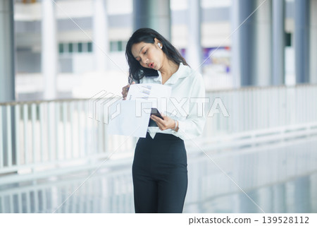 Professional businesswoman reviews financial charts on a clipboard in a modern office corridor, holding documents for corporate planning, data analysis, productivity, and business workflow concepts. Professional businesswoman reviews financial charts on a clipboard in a modern office corridor, holding documents for corporate planning, data analysis, productivity, and business workflow concepts. 139528112