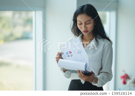 Professional businesswoman talks on a smartphone while holding reports near a bright office window, showing client communication, corporate coordination, mobile workflow, planning, and productivity. Professional businesswoman talks on a smartphone while holding reports near a bright office window, showing client communication, corporate coordination, mobile workflow, planning, and productivity. 139528114