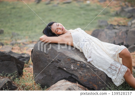 Woman in white lace dress lying peacefully on a large rock with eyes closed, soft outdoor nature portrait, bohemian summer fashion, relaxation, wellness lifestyle, restful travel, and calm beauty. Woman in white lace dress lying peacefully on a large rock with eyes closed, soft outdoor nature portrait, bohemian summer fashion, relaxation, wellness lifestyle, restful travel, and calm beauty. 139528157