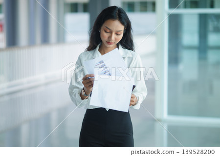 Focused businesswoman writes notes on a clipboard while leaning on a railing in a modern office corridor, representing financial review, administration, planning, and workplace productivity. Focused businesswoman writes notes on a clipboard while leaning on a railing in a modern office corridor, representing financial review, administration, planning, and workplace productivity. 139528200