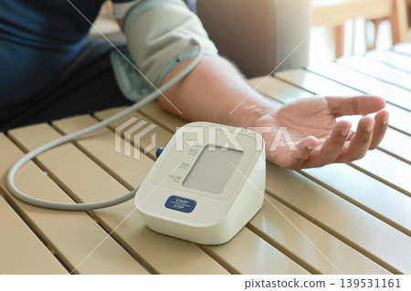 Unrecognizable man measuring blood pressure with digital sphygmomanometer at home. Close up of arm and automatic monitor on table. Healthcare, hypertension awareness and medical self care concept. 139531161