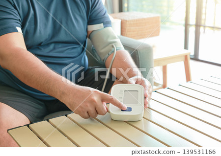 Unrecognizable overweight man pressing start button on digital blood pressure monitor at home. Obese male patient checking hypertension with automatic sphygmomanometer. Medical self care concept. Unrecognizable overweight man pressing start button on digital blood pressure monitor at home. Obese male patient checking hypertension with automatic sphygmomanometer. Medical self care concept. 139531166
