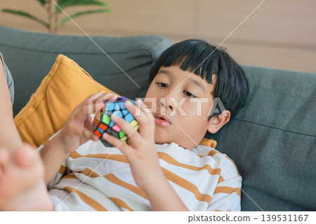 Smart Asian kid lying on a comfortable gray sofa, concentrating on solving a colorful dodecahedron puzzle toy. Child brain development, logical thinking, education and creative leisure time at home. Smart Asian kid lying on a comfortable gray sofa, concentrating on solving a colorful dodecahedron puzzle toy. Child brain development, logical thinking, education and creative leisure time at home. 139531167
