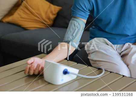 Asian woman measuring blood pressure with digital monitor at home. Female patient checking heart rate on sofa in living room with Christmas tree background. Healthcare and medical concept. Asian woman measuring blood pressure with digital monitor at home. Female patient checking heart rate on sofa in living room with Christmas tree background. Healthcare and medical concept. 139531168