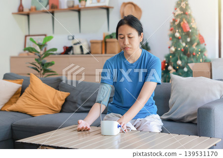 Serious Asian woman sitting on a sofa at home checking her blood pressure with a digital monitor during Christmas holidays. Healthcare, cardiology medical concept, hypertension wellness check. Serious Asian woman sitting on a sofa at home checking her blood pressure with a digital monitor during Christmas holidays. Healthcare, cardiology medical concept, hypertension wellness check. 139531170