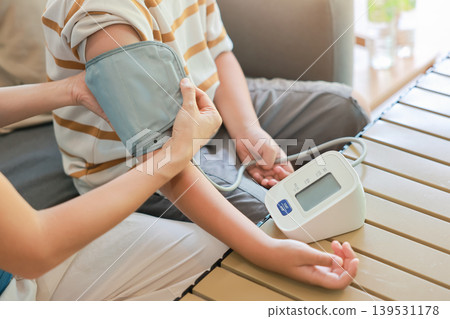 Close up unrecognizable mother hands applying blood pressure cuff on her son arm for measuring with automatic digital monitor at home. Family pediatric healthcare and daily wellness medical Close up unrecognizable mother hands applying blood pressure cuff on her son arm for measuring with automatic digital monitor at home. Family pediatric healthcare and daily wellness medical 139531178