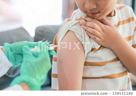 Close up of young Asian boy in a striped shirt receiving a vaccination injection shot in his upper arm from a doctor with green gloved hand. Medical checkup, pediatric immunization, virus prevention. 139531180