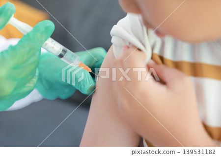 Close up of young Asian boy in a striped shirt receiving a vaccination injection shot in his upper arm from a doctor with green gloved hand. Medical checkup, pediatric immunization, virus prevention. Close up of young Asian boy in a striped shirt receiving a vaccination injection shot in his upper arm from a doctor with green gloved hand. Medical checkup, pediatric immunization, virus prevention. 139531182
