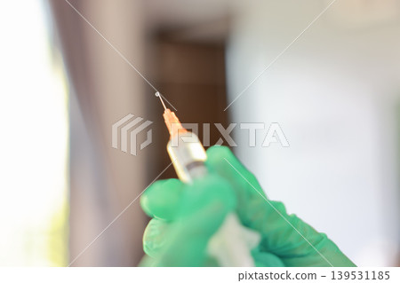 Close up macro shot of a sharp syringe needle tip with a small liquid drop. Unrecognizable doctor hand in a green latex glove preparing medicine vaccine dose for injection. Medical healthcare concept. 139531185