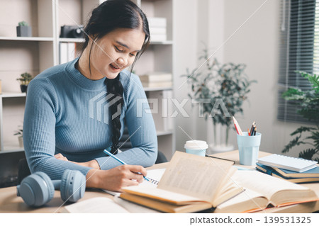 Young Asian woman writing study notes at home desk with open books, coffee and headphones nearby. Focused self study, exam preparation, reading, education and productivity concept. Young Asian woman writing study notes at home desk with open books, coffee and headphones nearby. Focused self study, exam preparation, reading, education and productivity concept. 139531325