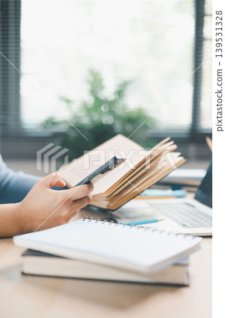 Young woman studying at home desk, holding smartphone and reading book beside laptop with headphones and coffee. Mobile learning, online research, digital education and study routine concept. Young woman studying at home desk, holding smartphone and reading book beside laptop with headphones and coffee. Mobile learning, online research, digital education and study routine concept. 139531328