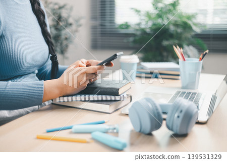 Young woman using smartphone over books beside laptop and headphones at home desk. Mobile learning, online study, digital research, multitasking, education and productivity concept. Young woman using smartphone over books beside laptop and headphones at home desk. Mobile learning, online study, digital research, multitasking, education and productivity concept. 139531329