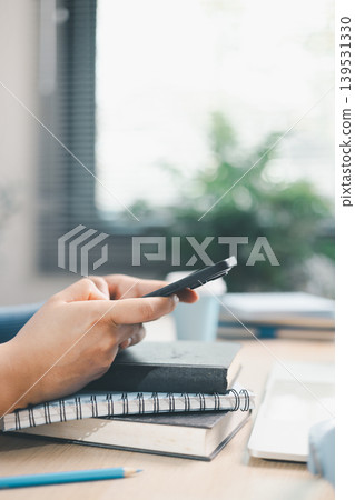 Young woman using smartphone over books beside laptop and headphones at home desk. Mobile learning, online study, digital research, multitasking, education and productivity concept. Young woman using smartphone over books beside laptop and headphones at home desk. Mobile learning, online study, digital research, multitasking, education and productivity concept. 139531330