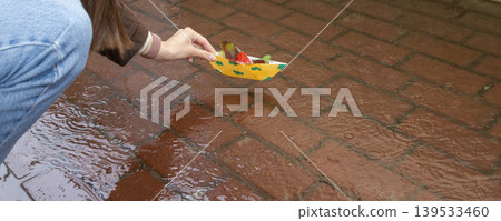 A person placing a yellow origami boat decorated with green clouds into a puddle on a red brick sidewalk A person placing a yellow origami boat decorated with green clouds into a puddle on a red brick sidewalk 139533460
