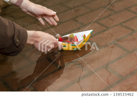 A person placing a yellow origami boat decorated with green clouds into a puddle on a red brick sidewalk A person placing a yellow origami boat decorated with green clouds into a puddle on a red brick sidewalk 139533462