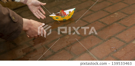 A person placing a yellow origami boat decorated with green clouds into a puddle on a red brick sidewalk A person placing a yellow origami boat decorated with green clouds into a puddle on a red brick sidewalk 139533463