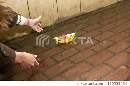 A person placing a yellow origami boat decorated with green clouds into a puddle on a red brick sidewalk A person placing a yellow origami boat decorated with green clouds into a puddle on a red brick sidewalk 139533464