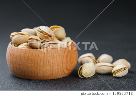 Little wooden bowl with salted pistachio nuts and scattered seeds on black background close up Little wooden bowl with salted pistachio nuts and scattered seeds on black background close up 139533718