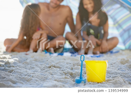 Family, sand and relax on beach with bucket, spade and umbrella for playing by ocean coast. Blue and yellow toy on sandy shore with people in fun bonding, shade or outdoor holiday weekend together Family, sand and relax on beach with bucket, spade and umbrella for playing by ocean coast. Blue and yellow toy on sandy shore with people in fun bonding, shade or outdoor holiday weekend together 139534999