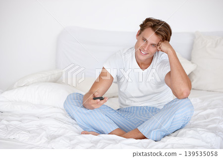 Happy man, morning and watching tv in bedroom with remote for streaming entertainment to relax. Smile, pyjamas or person enjoying television series, news or film with control for movie choice in home 139535058