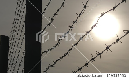 The barbed wires on the barrier of the guarded land from the coastal area. View from bottom to top. The sun is shining through the barbed wire. The barbed wires on the barrier of the guarded land from the coastal area. View from bottom to top. The sun is shining through the barbed wire. 139535660