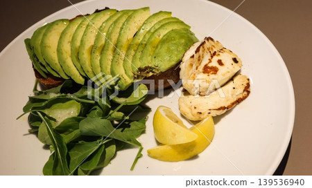 close up of avocado sourdough toast topped with sliced avocado. Baby rocket salad and grilled halloumi, added honey and lemon wedges served on the side, on a white plate with copy space close up of avocado sourdough toast topped with sliced avocado. Baby rocket salad and grilled halloumi, added honey and lemon wedges served on the side, on a white plate with copy space 139536940