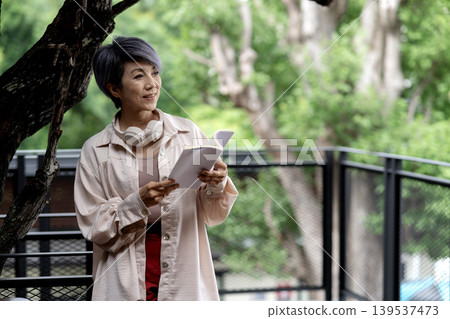 elder 50+ woman stand relax read book outdoor green tree elder 50+ woman stand relax read book outdoor green tree 139537473
