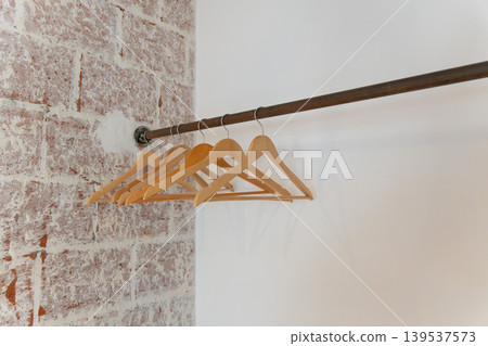 Wooden clothes hangers hang on metal rail against white wall, exposed brick, creating minimal interior detail with clean boutique mood. Retail design, closet organization, home staging, hospitality Wooden clothes hangers hang on metal rail against white wall, exposed brick, creating minimal interior detail with clean boutique mood. Retail design, closet organization, home staging, hospitality 139537573