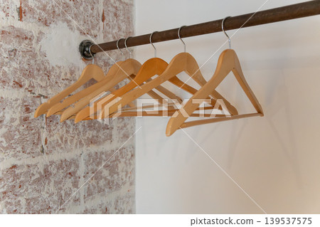 Wooden clothes hangers hang on metal rail against white wall, exposed brick, creating minimal interior detail with clean boutique mood. Retail design, closet organization, home staging, hospitality Wooden clothes hangers hang on metal rail against white wall, exposed brick, creating minimal interior detail with clean boutique mood. Retail design, closet organization, home staging, hospitality 139537575