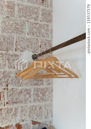Wooden clothes hangers hang on metal rail against white wall, exposed brick, creating minimal interior detail with clean boutique mood. Retail design, closet organization, home staging, hospitality Wooden clothes hangers hang on metal rail against white wall, exposed brick, creating minimal interior detail with clean boutique mood. Retail design, closet organization, home staging, hospitality 139537576