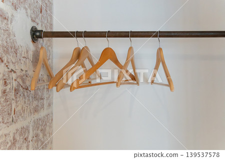 Wooden clothes hangers hang on metal rail against white wall, exposed brick, creating minimal interior detail with clean boutique mood. Retail design, closet organization, home staging, hospitality Wooden clothes hangers hang on metal rail against white wall, exposed brick, creating minimal interior detail with clean boutique mood. Retail design, closet organization, home staging, hospitality 139537578
