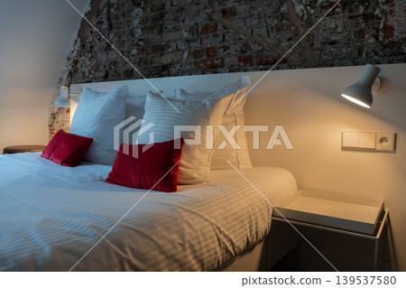 Hotel bed with white linen, pillows, red cushions, bedside table, wall lamp, and exposed brick wall creates cozy boutique room interior. Hospitality, travel accommodation, bedroom design, interior Hotel bed with white linen, pillows, red cushions, bedside table, wall lamp, and exposed brick wall creates cozy boutique room interior. Hospitality, travel accommodation, bedroom design, interior 139537580