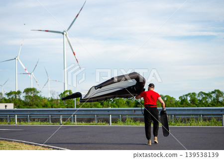 Skateboarder with wing foil on empty road near wind farm Skateboarder with wing foil on empty road near wind farm 139538139