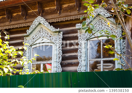Intricately carved windows adorn an old wooden log house, showcasing traditional Russian craftsmanship and heritage, partially obscured by lush green foliage 139538150
