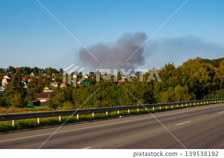 Thick grey smoke billows above a village with homes nestled among lush trees, creating an alarming scene beside a quiet road. A potential danger or emergency unfolding Thick grey smoke billows above a village with homes nestled among lush trees, creating an alarming scene beside a quiet road. A potential danger or emergency unfolding 139538192