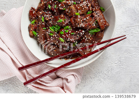 Crispy beef, with green onions and sesame seeds, in a bowl with chopsticks, on a textured surface, on a light background, no people 139538679