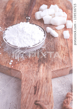 a strainer with powdered sugar, on a wooden cutting board, with pieces of white sugar, 139538715