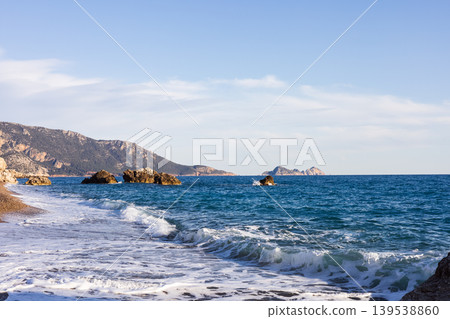 Blue sea waves crashing on pebble beach with distant mountains 139538860