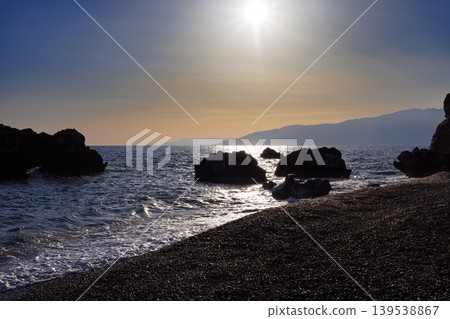 Sunlit rocky coastline with calm waves at sunset Sunlit rocky coastline with calm waves at sunset 139538867