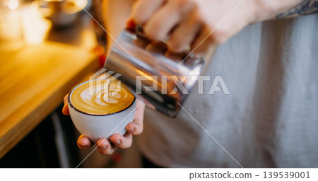 Cappuccino preparation. Hot beverage. Man hand pouring steamed milk from steel jug in espresso coffee mug making white foam pattern. Cappuccino preparation. Hot beverage. Man hand pouring steamed milk from steel jug in espresso coffee mug making white foam pattern. 139539001
