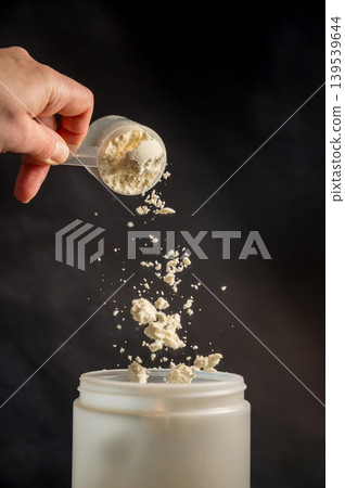 Protein drink. Hand pouring protein powder in a white shaker. Dark background. Body building, muscle growth and food supplement concept Protein drink. Hand pouring protein powder in a white shaker. Dark background. Body building, muscle growth and food supplement concept 139539644