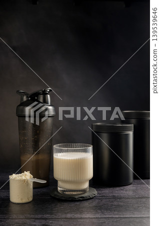 Protein drink in a glass with protein shaker and tube on dark background. Body building, muscle growth and food supplement concept Protein drink in a glass with protein shaker and tube on dark background. Body building, muscle growth and food supplement concept 139539646