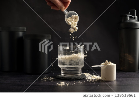 Protein drink. Hand pouring protein powder in a glass of water. Dark background. Body building, muscle growth and food supplement concept Protein drink. Hand pouring protein powder in a glass of water. Dark background. Body building, muscle growth and food supplement concept 139539661